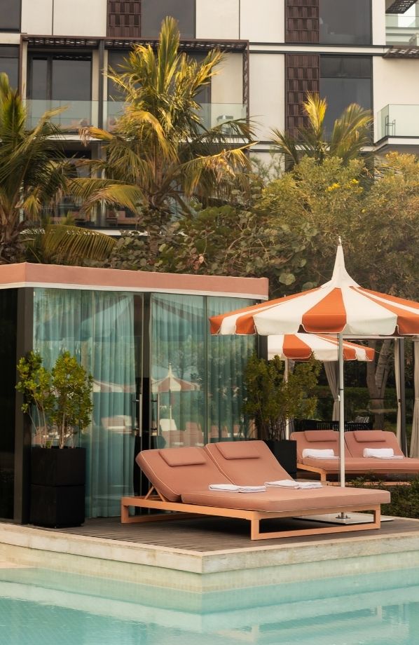 Elegant poolside setting featuring plush double sun loungers with soft pink cushions, neatly placed towels, and an orange-and-white striped umbrella. A private cabana with floor-to-ceiling glass doors and sheer curtains sits behind, surrounded by lush greenery. The modern architectural backdrop includes glass balconies and decorative wooden panels, enhancing the luxurious resort ambiance.